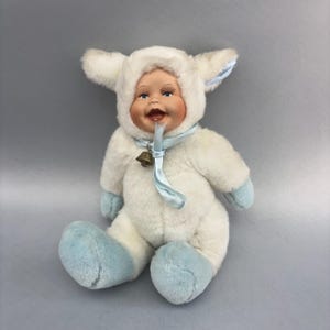 May include: A plush doll featuring a porcelain-like face with blue eyes and an open mouth, wearing a white fuzzy hood with animal ears. The doll has a light blue ribbon tied under the chin with a small metal bell and light blue plush feet.