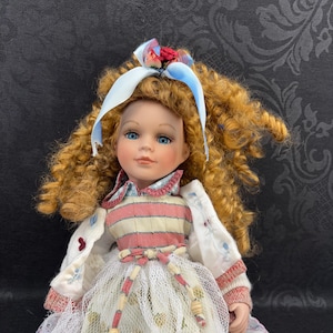 May include: A porcelain doll with curly ginger hair, blue eyes, and a light complexion. The doll wears a white cardigan with red trim over a pink and white striped bodice, a white lace skirt, and a blue ribbon with small red flowers in its hair.