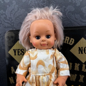 Op de afbeelding: Een vintage pop met een perzikkleurig gezicht, blauwe ogen en lichtroze haar. De pop draagt een witte jurk met een herhalend oranje patroon. Een zwart Ouija-bord is op de achtergrond zichtbaar met het woord "YES".