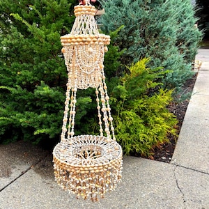 May include: A hanging chandelier-style decorative piece crafted from numerous small, light-coloured seashells. The structure features a tiered design with a cylindrical top section and a wider, circular base, both adorned with cascading shell strands.