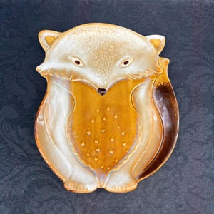 May include: A ceramic dish shaped like a fox, featuring a glossy finish in shades of golden brown and cream. The fox design includes embossed details on the chest, pointed ears, and a curved tail, creating a functional tray with a smooth surface.