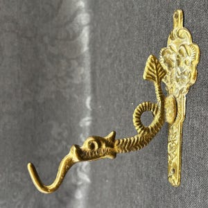 Può includere: Un gancio da parete decorativo in ottone con un design a drago. Il gancio ha un'estremità curva e aperta e una testa di drago dettagliata. La piastra di montaggio è ornata con motivi floreali. Il gancio è di colore oro caldo.
