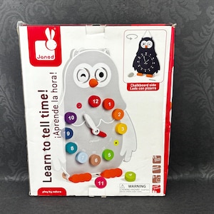 May include: A boxed owl-shaped wooden clock toy with colorful hour markers. The clock face has a white background with a clock hand. The box has the text "Learn to tell time!" and "play by nature".