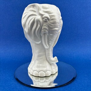 May include: White ceramic elephant head vase. The vase is detailed with textured ears, a trunk, and feet. The vase is sitting on a silver mirrored surface.