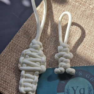 Puede incluir: Dos llaveros de cuerda blanca con un diseño de nudo. El llavero más grande tiene un diseño de nudo más complejo. El llavero más pequeño tiene un diseño de nudo más simple. Ambos llaveros están sobre un fondo de arpillera.