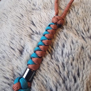 Puede incluir: Un llavero de paracord trenzado de color azul verdoso y marrón con una cuenta de metal negro. El llavero tiene un bucle en la parte superior para sujetarlo a las llaves o a una bolsa.