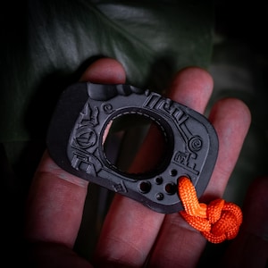 May include: A matte black, rectangular-shaped EDC tool featuring a central circular cutout, intricate engraved geometric patterns on the surface, and a bright orange paracord lanyard attached through a small hole at the bottom corner.