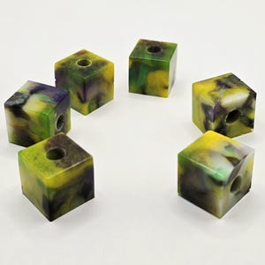 以下が含まれることがあります： 中央に穴の開いた、正方形の樹脂ビーズが6つ。ビーズは黄色、緑、紫の色が混ざっています。ビーズは白い表面に円を描くように配置されています。これらのビーズは、ジュエリー作りやその他のクラフトプロジェクトに使用できます。