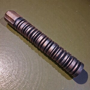 Könnte beinhalten: Ein bronzefarbenes, zylindrisches Objekt mit einem geriffelten, spiralförmigen Design. Die Oberseite hat eine flache, geometrische Form. Das Objekt ist metallisch und scheint aus Metall zu sein. Der Artikel liegt auf einer grünen Oberfläche.