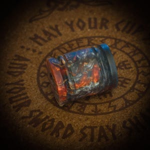 May include: A cylindrical bead featuring a marbled pattern of deep orange, charcoal grey, and black swirls. The bead has a smooth, polished finish with a distinct grooved texture around its circumference, creating a multi-toned appearance.