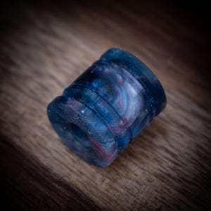 May include: A cylindrical bead featuring a marbled pattern of deep blue, navy, and subtle hints of reddish-purple. The bead has a smooth, polished finish with two distinct horizontal grooves encircling its exterior and a central hole.