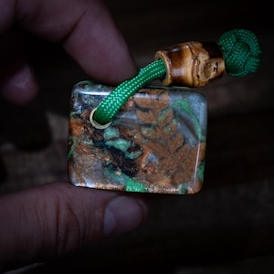Peut inclure: Un pendentif rectangulaire en résine intégrant une feuille de fougère brune et des flocons métalliques cuivrés avec des accents verts. Une boucle en paracorde vert vif traverse un trou dans la résine, fixée par une perle en bois et un nœud.