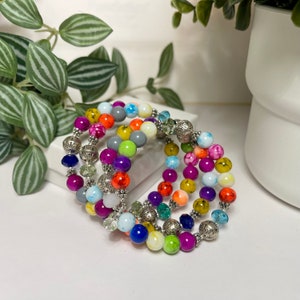 May include: A set of four colorful beaded bracelets with silver accents. The bracelets feature a variety of bright colors, including pink, blue, green, yellow, and orange. The beads are made of glass and have a slightly iridescent finish.