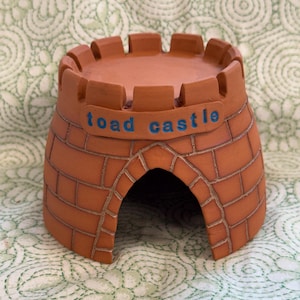 May include: A terracotta-colored ceramic toad house shaped like a castle with a crenelated top and a brick pattern etched into the exterior. The front features a prominent arched doorway and a raised rectangular plaque with the text toad castle.