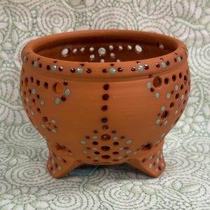 Può includere: Un vaso di terracotta a tre gambe con fori decorativi e accenti dipinti. Il vaso presenta una serie di piccoli fori rotondi ed è adornato da punti turchese e marroni. Il bordo è anch'esso decorato con punti turchese.
