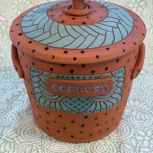 May include: A handmade terracotta compost bin with a lid and handle. The bin has a turquoise and brown design and the word "COMPOST" on the front. The lid and body are decorated with small, dark brown dots.
