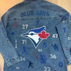 Peut inclure: Veste en jean avec l'inscription "BLUE JAYS" en lettres bleues pailletées au dos. La veste présente un logo des Blue Jays, des chiffres et des graphiques sur le thème du baseball.
