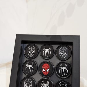 May include: A black framed display with nine round black and silver Spiderman themed buttons. The buttons feature a variety of Spiderman images, including the classic red and blue suit, a black suit, and a spider web.