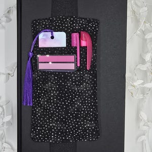 May include: A black journal with a black elastic band and a pocket with a black and white polka dot pattern. The pocket holds a pink pen, a pink highlighter, a pink and white tag, and two pink rectangular sticky notes. A purple tassel hangs from the tag.
