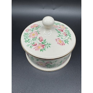 May include: A round, off-white ceramic trinket box with a matching lid featuring a knob handle. The surface is decorated with a pattern of pink flowers and green leaves, accented by a thin gold trim along the edges of the lid and the base.