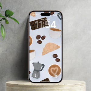 May include: Smartphone screen displaying a coffee-themed wallpaper. The design features coffee cups, beans, a croissant, chocolate, and a coffee maker. The time is 11:54 on Monday, December 22. The phone is on a wooden stand.
