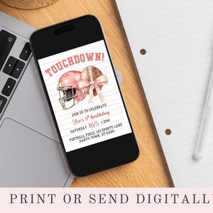 May include: A smartphone displays a pink football-themed birthday invitation. The invitation features a pink football helmet and football with a bow, and the text "TOUCHDOWN!" The phone rests on a wooden surface next to a laptop and a notepad.