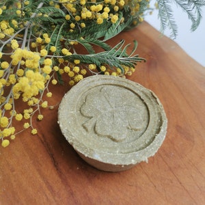Peut inclure: Un savon rond et vert avec un motif de trèfle, posé sur une surface en bois. Des fleurs de mimosa jaunes et du feuillage vert en arrière-plan ajoutent une touche naturelle à la composition.