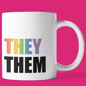 May include: A white ceramic mug featuring the word THEY in a vertical rainbow gradient font and the word THEM in bold black capital letters. The mug has a standard cylindrical shape with a curved handle on the right side, presented clearly.