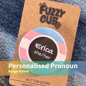 May include: A circular 45mm button badge featuring a light blue, pink, and white striped background with a central black circle containing the white text Erica and she/her, mounted on a brown cardboard display card labelled Fuzzy Cub.