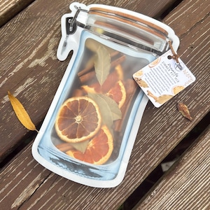 May include: A clear, jar-shaped pouch filled with dried orange slices, cinnamon sticks, and bay leaves. The pouch has a wooden lid and a metal clasp. A tag reads "Prosperity Simmer Pot Mix."