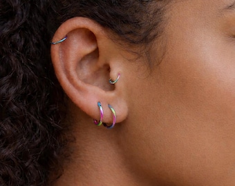 May include: Close-up of an ear adorned with multiple hoop earrings. The earrings display a vibrant, iridescent color scheme, transitioning between shades of blue, green, and pink. The jewelry is a modern accessory.