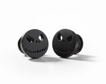 May include: Two black ear gauges featuring a carved design of a smiling face with oval eyes and a stitched mouth. The gauges are round and have a flat front with a slightly raised edge.