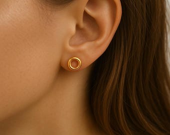 May include: A close-up of a person's ear with a gold-colored, circular stud earring. The earring has a hollow center and a smooth, polished finish. The earring is a simple, elegant design.