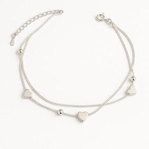 Op de afbeelding: Een zilverkleurige enkelband met een dubbelstrengs ontwerp. De enkelband is versierd met hartvormige bedels en kleine, ronde kralen. De verstelbare ketting zorgt voor een comfortabele pasvorm.