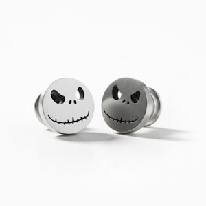Könnte beinhalten: Zwei silberfarbene Ohr-Dehnstäbe mit dem Gesicht von Jack Skellington aus Nightmare Before Christmas. Jeder Dehnstab hat ein kreisförmiges Gesicht mit ausgeschnittenen Augen und einem genähten Lächeln. Auf weißem Hintergrund.