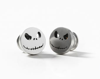 May include: Two silver-colored ear gauges featuring the face of Jack Skellington from The Nightmare Before Christmas. Each gauge has a circular face with cut-out eyes and a stitched smile. The gauges are displayed on a white background.