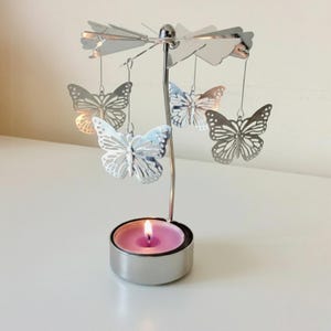 May include: A silver-colored candle holder with a rotating butterfly design. The candle holder has a round base with a pink candle inside. Five butterfly ornaments hang from the top, and the candle is lit.