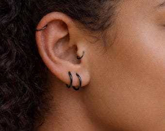May include: Close-up of an ear with multiple black hoop earrings. The earrings vary in size and are placed in different piercings. The skin tone is a warm brown, and the hair is dark and curly.