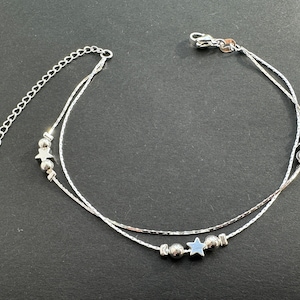 Może przedstawiać: Srebrna bransoletka na kostkę o podwójnym łańcuszku. Bransoletka ma małe zawieszki w kształcie gwiazd i okrągłe koraliki. Posiada regulowany łańcuszek. Biżuteria jest prezentowana na ciemnej powierzchni.