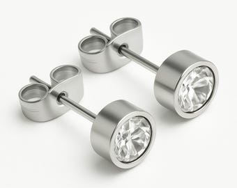 May include: A pair of silver-colored stud earrings. Each earring features a round, bezel-set, clear crystal. The crystals are surrounded by a polished, metallic rim. The earring posts and backs are also silver-colored.