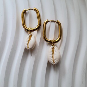 Op de afbeelding: Een paar gouden oorringen met een witte schelpbedel die aan elke oorring hangt.
