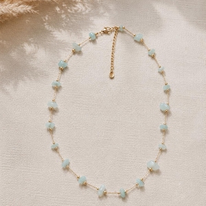 Könnte beinhalten: Eine zarte, goldfarbene Halskette mit kleinen, hellblauen Edelsteinperlen. Die Halskette hat eine feine Kette mit gleichmäßig verteilten Edelsteinen und kleinen goldenen Perlen. Der Verschluss und die Kette sind sichtbar.