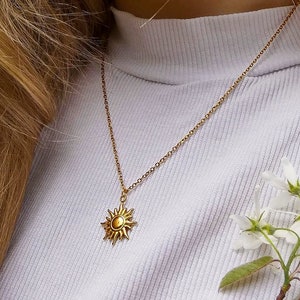Op de afbeelding: Een gouden ketting met een zon hanger. De ketting wordt gedragen door een persoon met lang blond haar. De zon hanger heeft een gedetailleerd ontwerp met stralen.