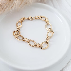 Könnte beinhalten: Ein goldfarbenes Kettenarmband mit ovalen Gliedern und Knotendetails. Das Armband wird auf einem weißen Teller vor einem weichen, neutralen Hintergrund präsentiert. Mit Karabinerverschluss.
