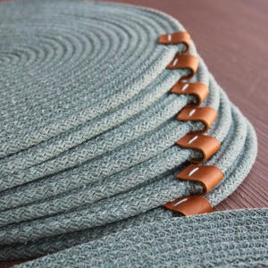 Peut inclure: Une pile de sets de table circulaires en corde tressée vert sauge, chacun présentant un motif en spirale texturé et une languette en cuir beige fixée sur le bord avec des coutures blanches. Les sets sont empilés pour montrer le design.