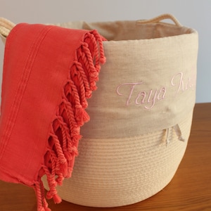 May include: A beige rope basket with a linen top and pink embroidery that says "Taya Rose". A coral coloured fringed towel is draped over the side of the basket.