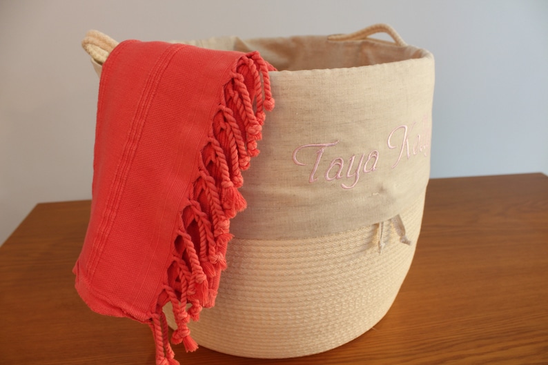 May include: A beige rope basket with a linen top and pink embroidery that says "Taya Rose". A coral coloured fringed towel is draped over the side of the basket.