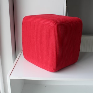 Puede incluir: Un puf de forma c&uacute;bica de color rojo brillante con una superficie de punto acanalado texturizado. El objeto tiene esquinas redondeadas y una forma s&oacute;lida y estructurada, presentando una apariencia uniforme en todos sus lados visibles.