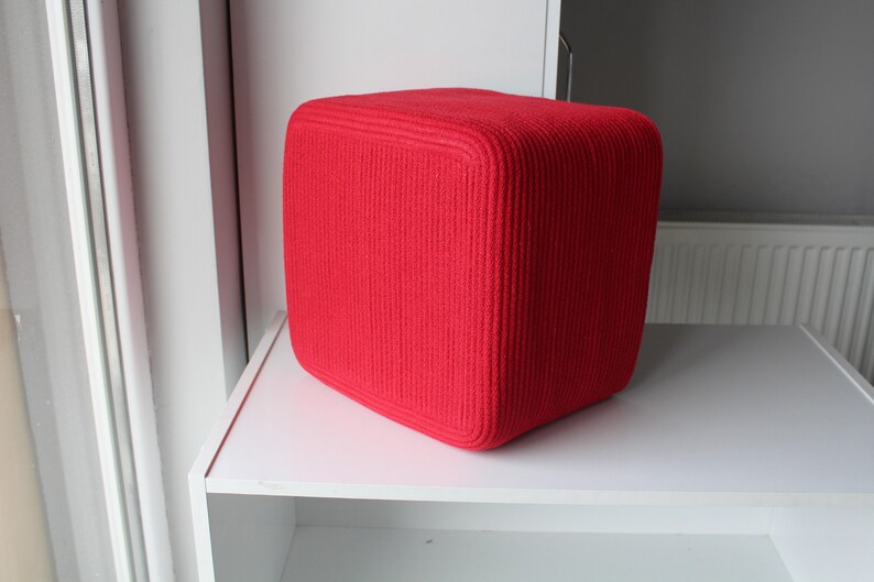 Puede incluir: Un puf de forma c&uacute;bica de color rojo brillante con una superficie de punto acanalado texturizado. El objeto tiene esquinas redondeadas y una forma s&oacute;lida y estructurada, presentando una apariencia uniforme en todos sus lados visibles.