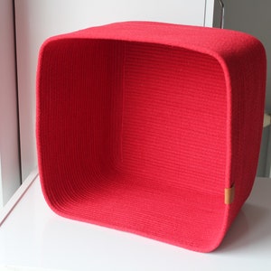 Puede incluir: Una cesta de almacenamiento cuadrada construida con cuerda roja enrollada. La cesta presenta esquinas redondeadas y una peque&ntilde;a leng&uuml;eta rectangular de color cuero tostado unida al lado inferior derecho de la pared exterior.
